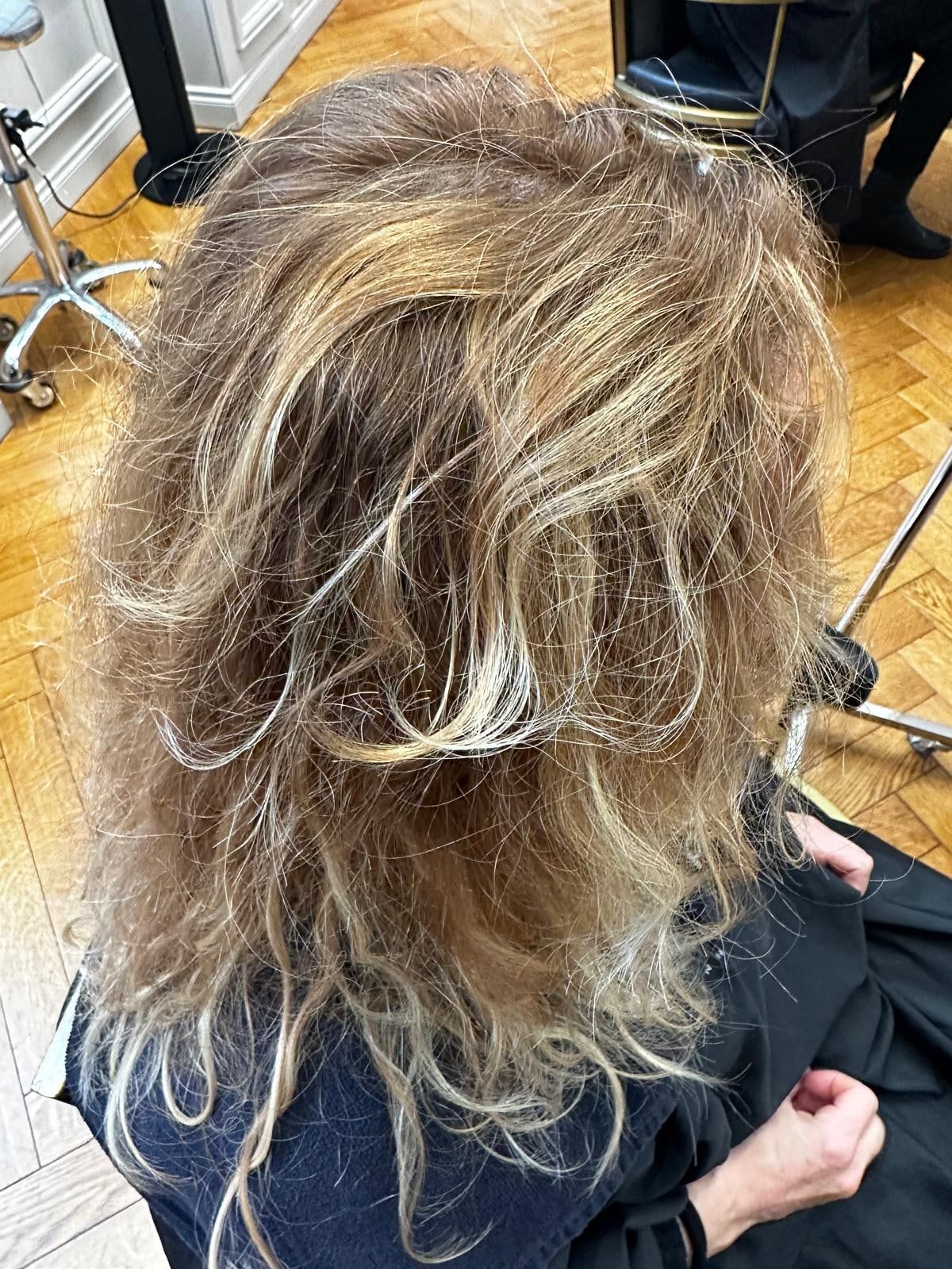 Avant le lissage : cheveux blonds méchés, secs et frisottants, vus de dos au salon sur le parquet à chevrons