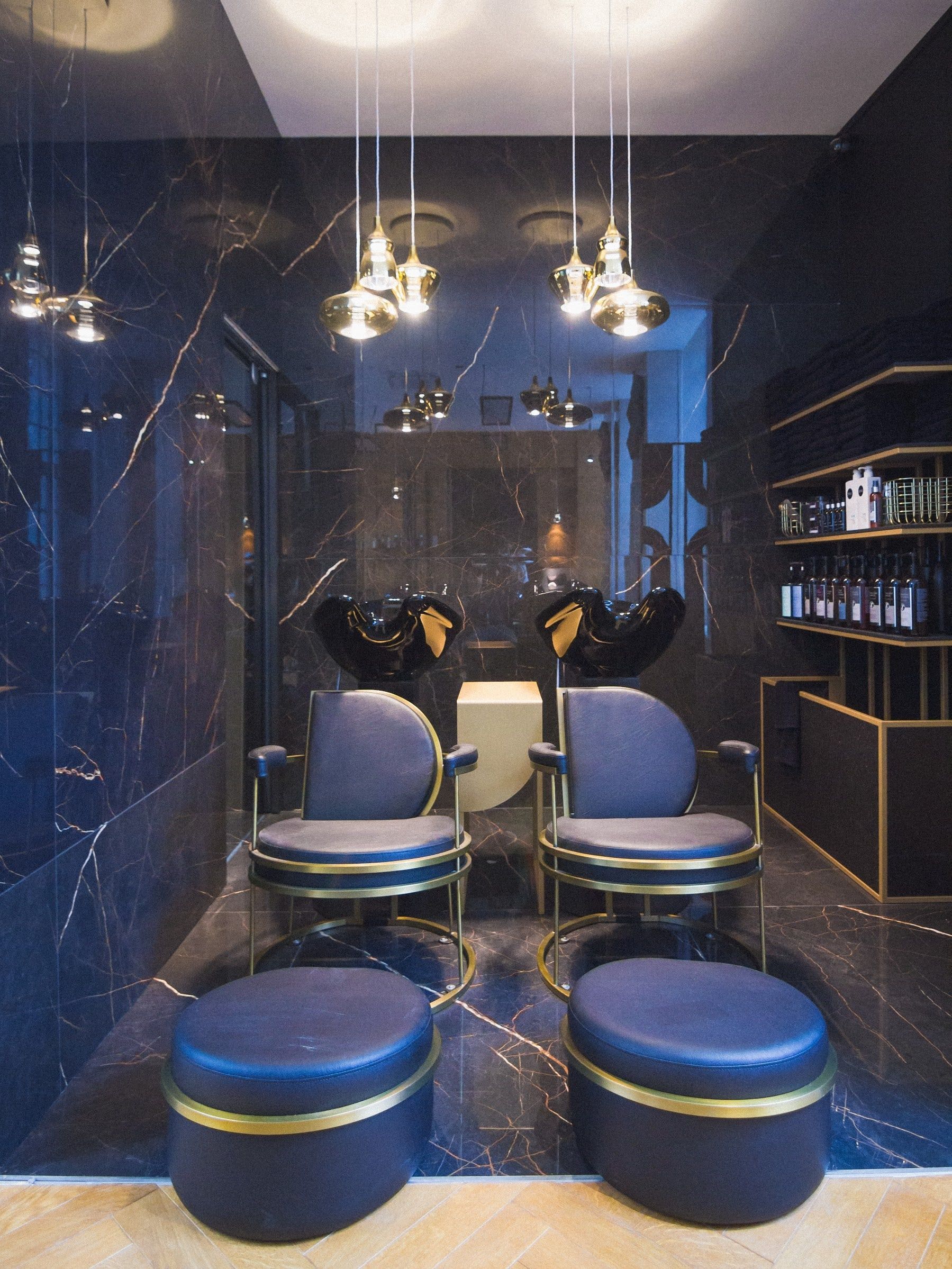 Espace shampoing en marbre noir veiné d'or, deux bacs avec fauteuils et repose-pieds bleu nuit cerclés de laiton, suspensions en verre ambré