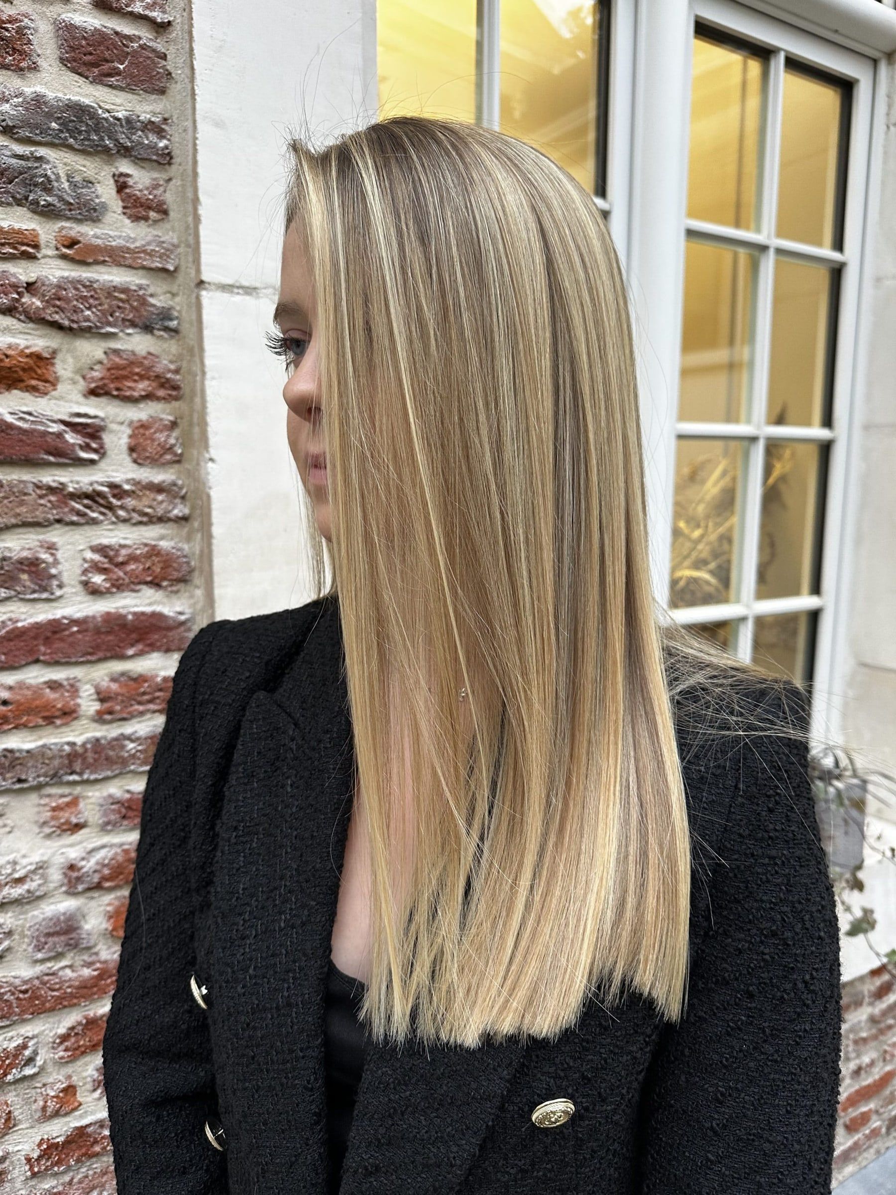 Cliente aux cheveux longs et lisses éclaircis d'un balayage blond miel, devant une façade en brique du Vieux-Lille