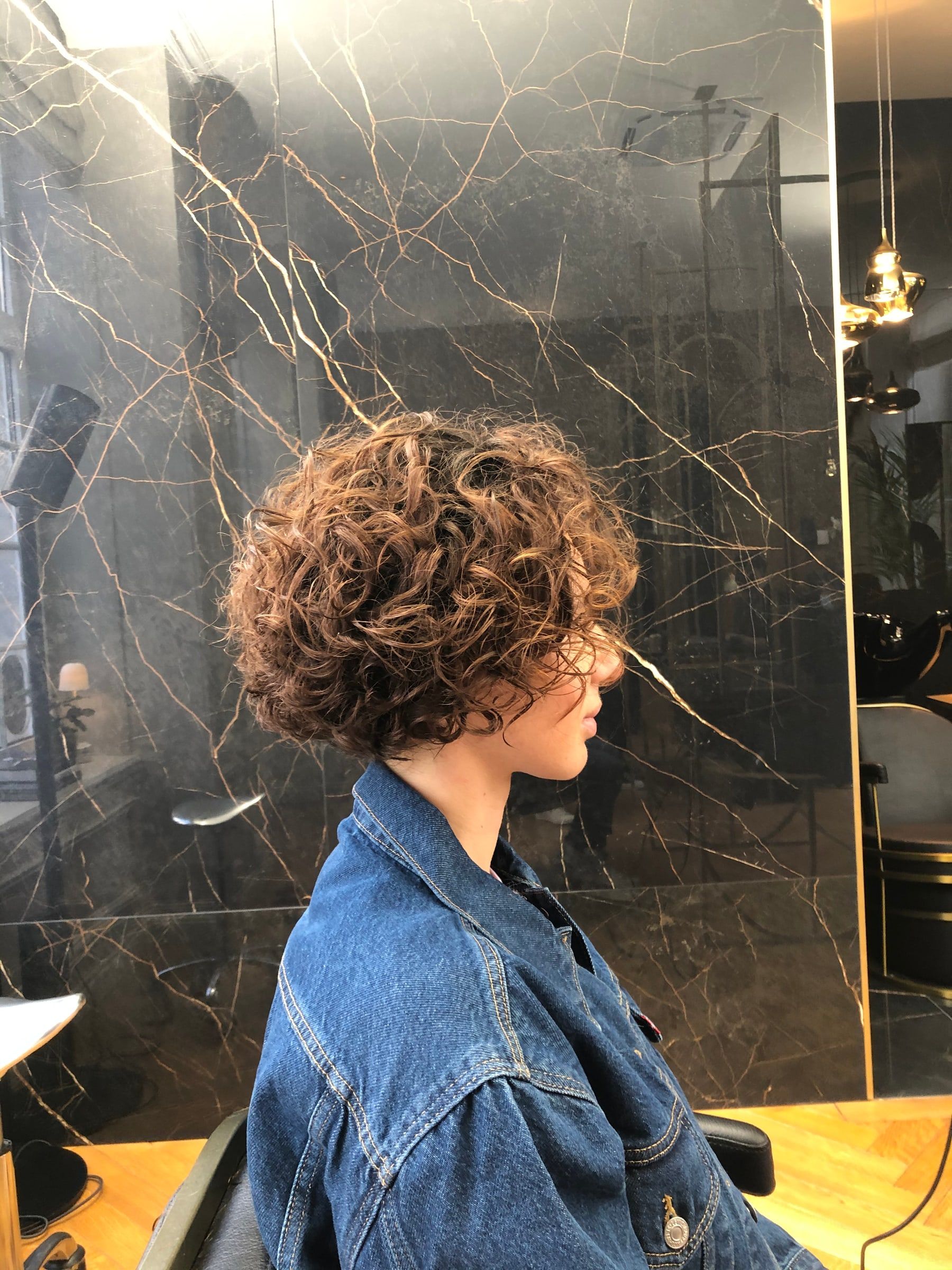 Coupe courte sur cheveux bouclés châtains, vue de profil devant le marbre noir du salon
