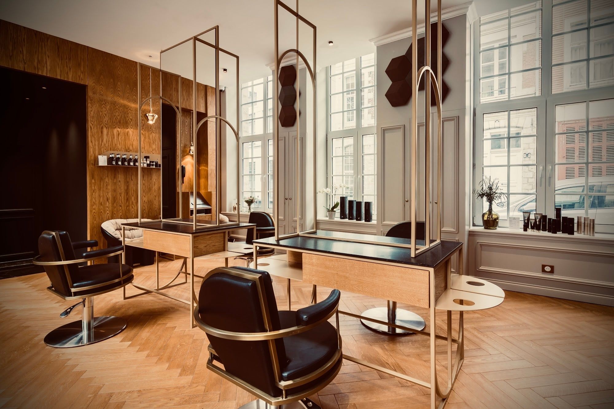 Espace coupe éclairé par de hautes fenêtres à petits carreaux, avec boiseries claires, miroirs en arche dorés et fauteuils en cuir