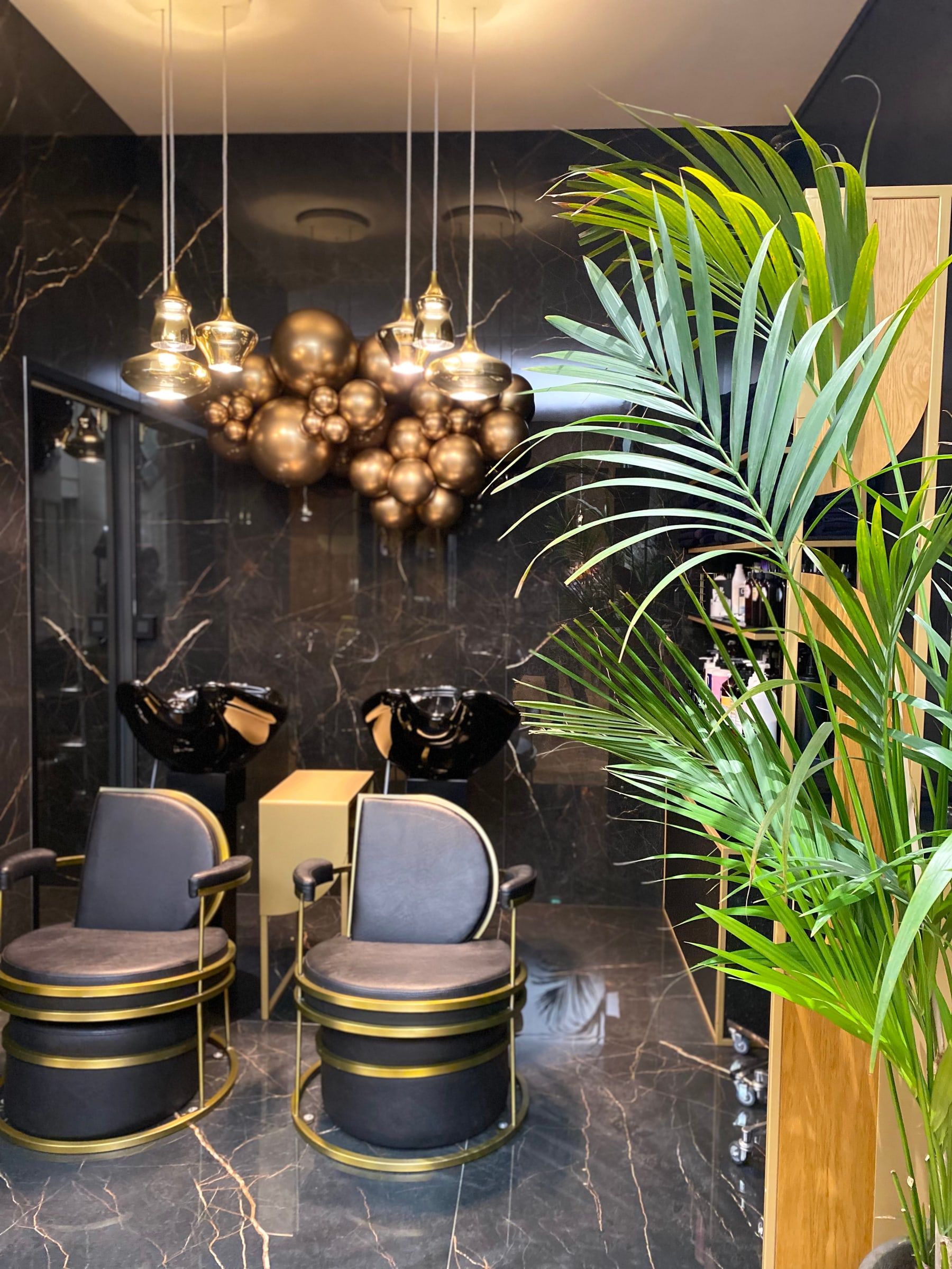 Bacs à shampoing sous un lustre de sphères dorées, murs en marbre sombre et grand palmier en pot
