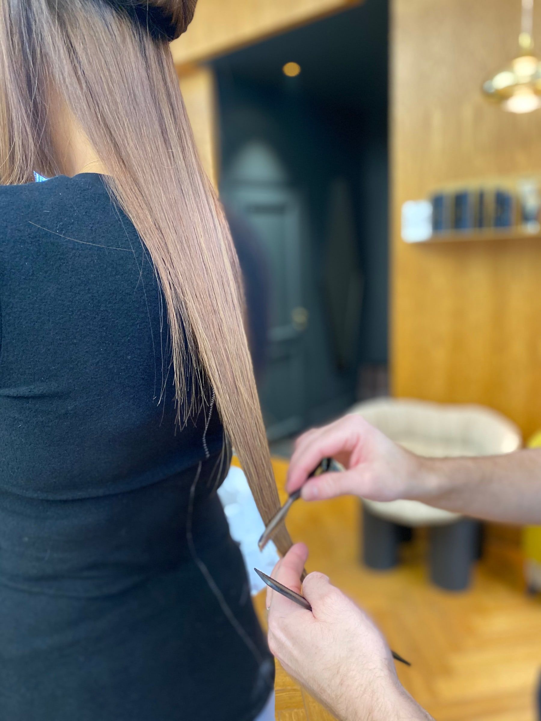 Coiffeur égalisant aux ciseaux et au peigne les longueurs d'une cliente après la pose d'extensions, dans le salon aux murs boisés