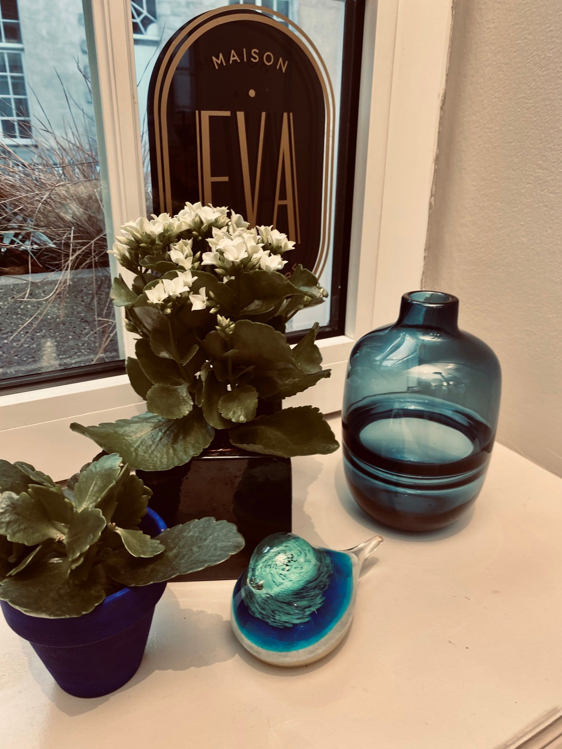 Rebord de fenêtre décoré de plantes fleuries, d'un vase en verre bleu et d'un oiseau en verre, devant le logo Maison de Coiffure Eva